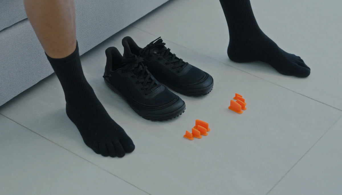 Black toe-socked feet beside black Arc trainers and two orange FeetFormers on a tiled floor.