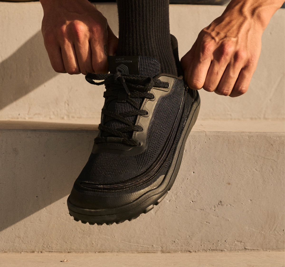 Hands adjusting the laces and side zipper of a black Arc trainer.